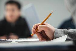 Hand holding a pencil over a notebook, out of focus student in the background.